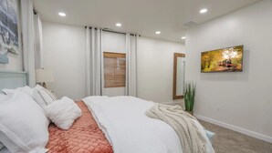 Une chambre à coucher comprenant un lit, une table de chevet, un miroir, une fenêtre avec des stores, un tableau et une plante en pot.