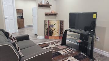 A living room with a gray sofa, a black entertainment unit with a TV, a patterned rug, and a kitchen in the background.