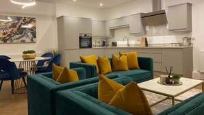 A modern kitchen with white cabinets, a built-in oven, and a microwave. There is a dining area with a table and chairs, and a sofa with yellow cushions. - A 2 bedroomed penthouse with large bi-fold doors accessing a good sized decked balcony area with stu (York)