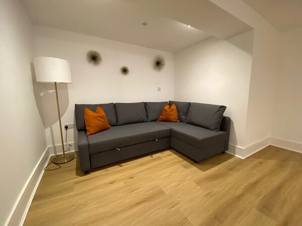 Un salon moderne avec un canapé d’angle gris, deux coussins orange, une lampe sur pied et des appliques murales décoratives.