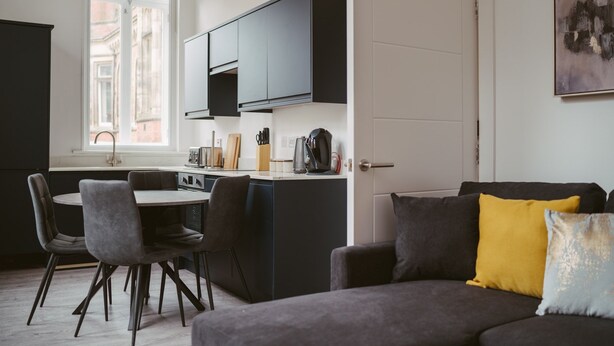 A modern kitchen with dark cabinets, a dining area with two chairs, and a sofa with a yellow pillow. - Apartment 3 (1 bedroom, sleeps x 4) (York)