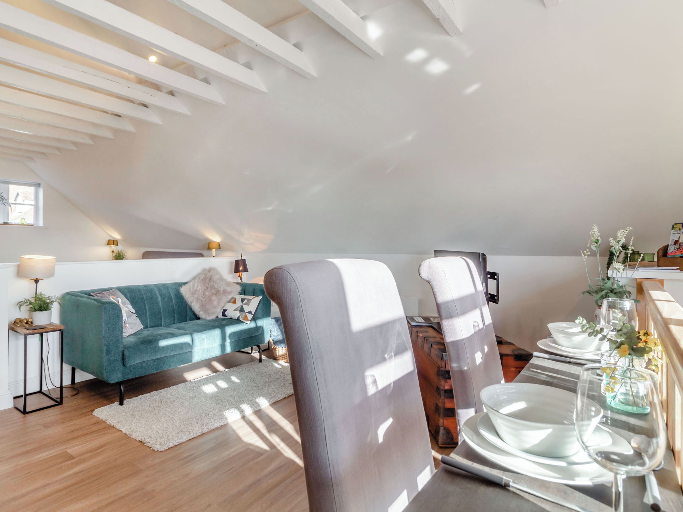 A modern loft bedroom with a sloped ceiling, a teal sofa, a dining table set with plates and glasses, and a small side table with a lamp.