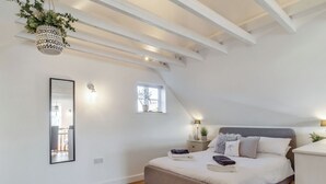 Une chambre avec un plafond en pente, un lit avec des oreillers et une couverture, une table de chevet avec une lampe, une plante suspendue et une fenêtre.