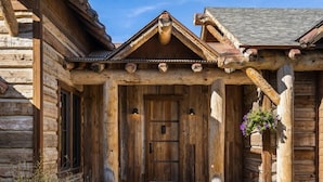 A rustic log cabin with a wooden front door, a porch with columns, and a hanging flower basket. - Stunning 5 Bedroom Big Sky Home on 20 Acres (Big Sky)
