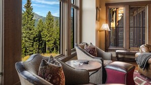A cozy room with a large window overlooking a forest and mountains. There are two leather armchairs and a small round table with a remote control. - Stunning 5 Bedroom Big Sky Home on 20 Acres (Big Sky)