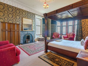 A four-poster bed, a fireplace, two armchairs, and a large mirror in a room with patterned wallpaper and a view of the outdoors.