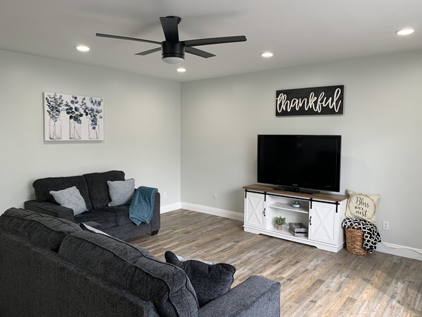 Ein modernes Wohnzimmer mit einem grauen Sofa, einem Flachbildfernseher auf einem Holzbodenständchen, einem Deckenventilator und einem Schild mit der Aufschrift 'thankful'.