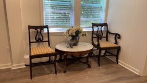 Een eetruimte met twee houten stoelen en een ronde tafel, een vaas met bloemen en een raam met zonwering.