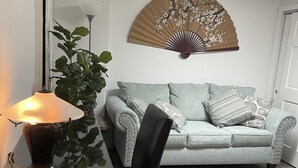 Een woonkamer met een lichtblauwe bank, een zwarte stoel, een tafel met een lamp en een decoratieve ventilator aan de muur.