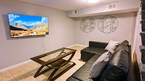 En stue med en sort lædersofa, et glasbord, et fladskærms-tv på væggen og dekorativ vægkunst.