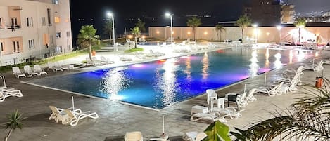 A nighttime swimming pool with lounge chairs and palm trees, surrounded by buildings.