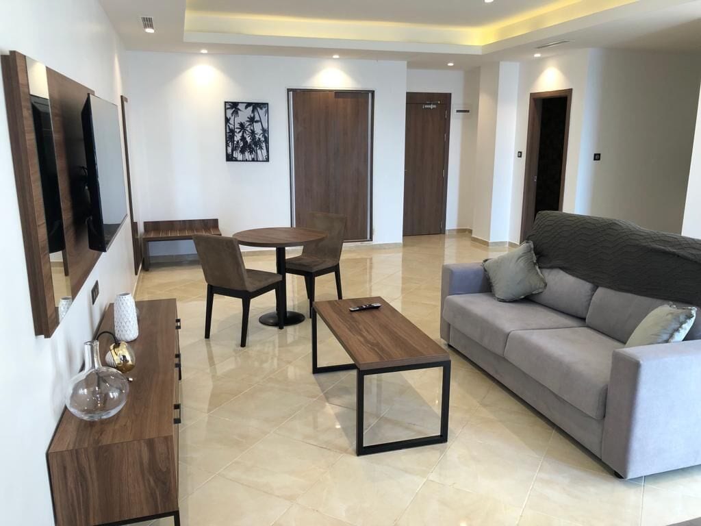A modern living room with a grey sofa, a wooden coffee table, a TV mounted on the wall, and a dining area with a round table and chairs.
