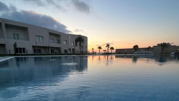 En swimmingpool med flisegulv, omgivet af en bygning med flere vinduer og altaner, under en delvist overskyet himmel.