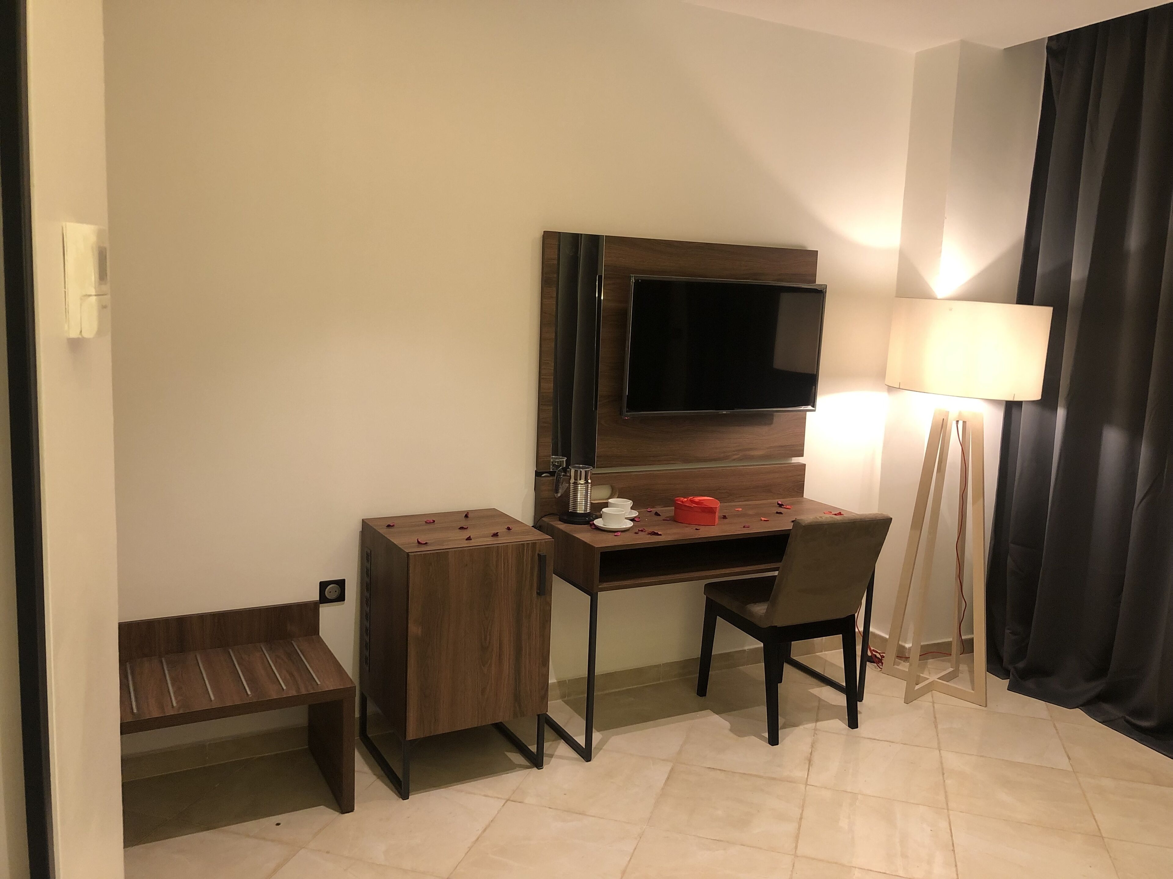Habitación de hotel con un escritorio de madera, una silla, un televisor montado en la pared, una lámpara de pie y un pequeño banco.