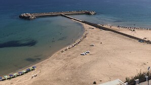 En strand med en buet mole, der strækker sig ud i havet, klare himmel og en sandet kystlinje med parasoller og strandstole.