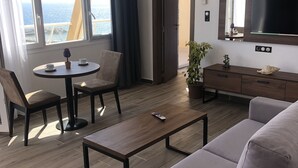 Et moderne hotelværelse med en sofa, et sofabord, et spisebord med stole, et fjernsyn og en balkon med havudsigt.