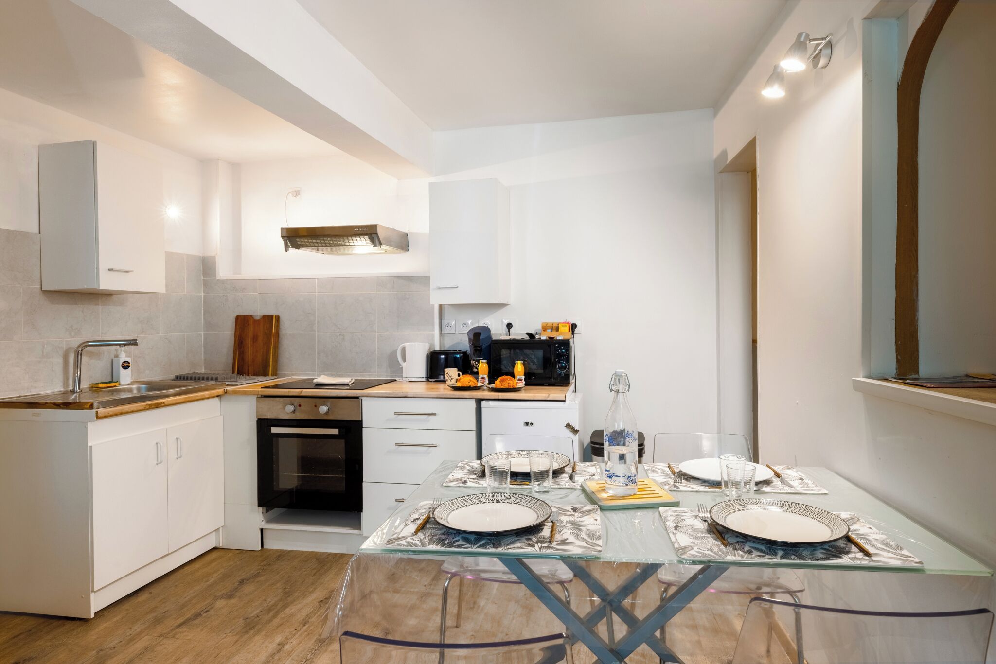 Une cuisine moderne avec des armoires blanches, un four encastré et une table à manger en verre pour quatre personnes.