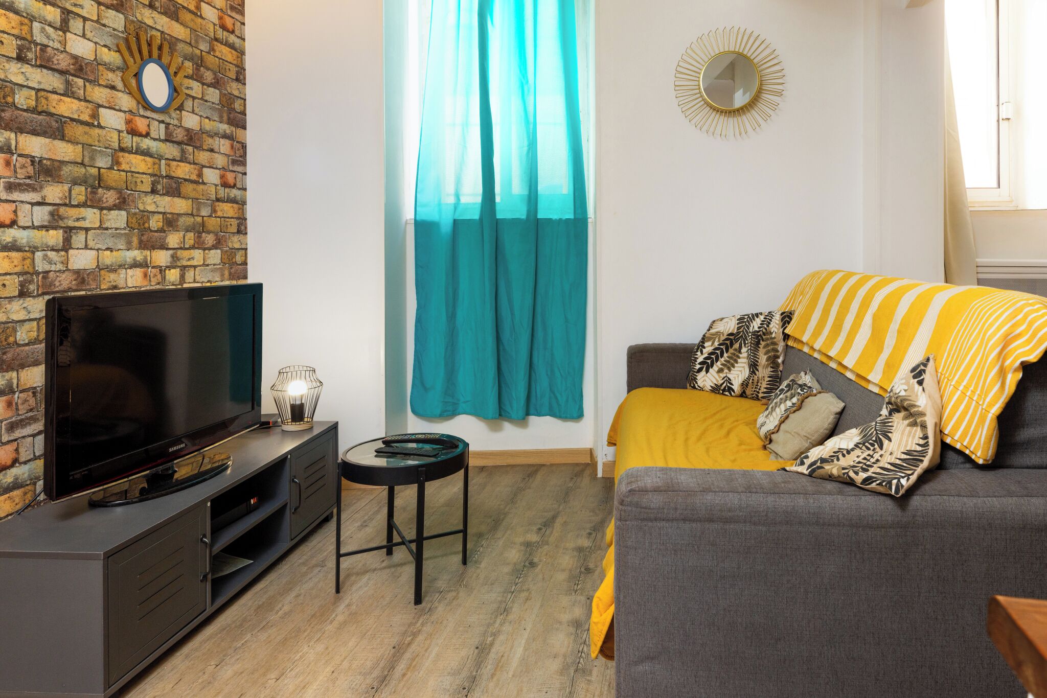 Un salon avec un téléviseur à écran plat posé sur un meuble en bois foncé, une petite table ronde avec une lampe, un canapé recouvert d’un plaid jaune et blanc à rayures, et un rideau de couleur turquoise.