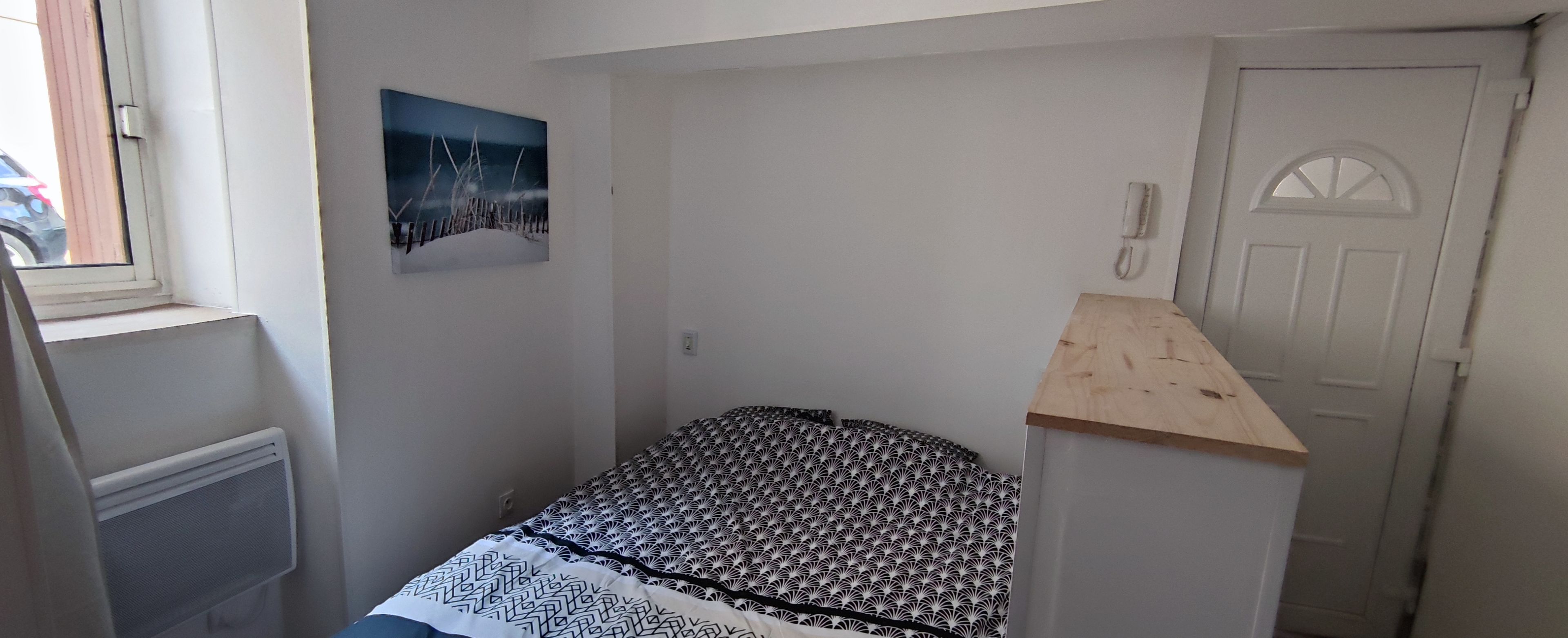 A small, single-bed room with a patterned bedspread, a radiator, a wall-mounted phone, and a door with a window.