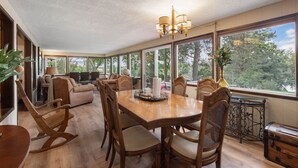 Un comedor amplio con una mesa y sillas de madera, ventanas grandes que ofrecen vistas a árboles y montañas, y una araña colgante.