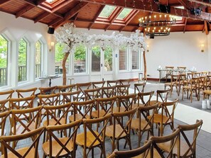 An indoor venue with wooden chairs arranged in rows, large windows, and a wooden ceiling structure.