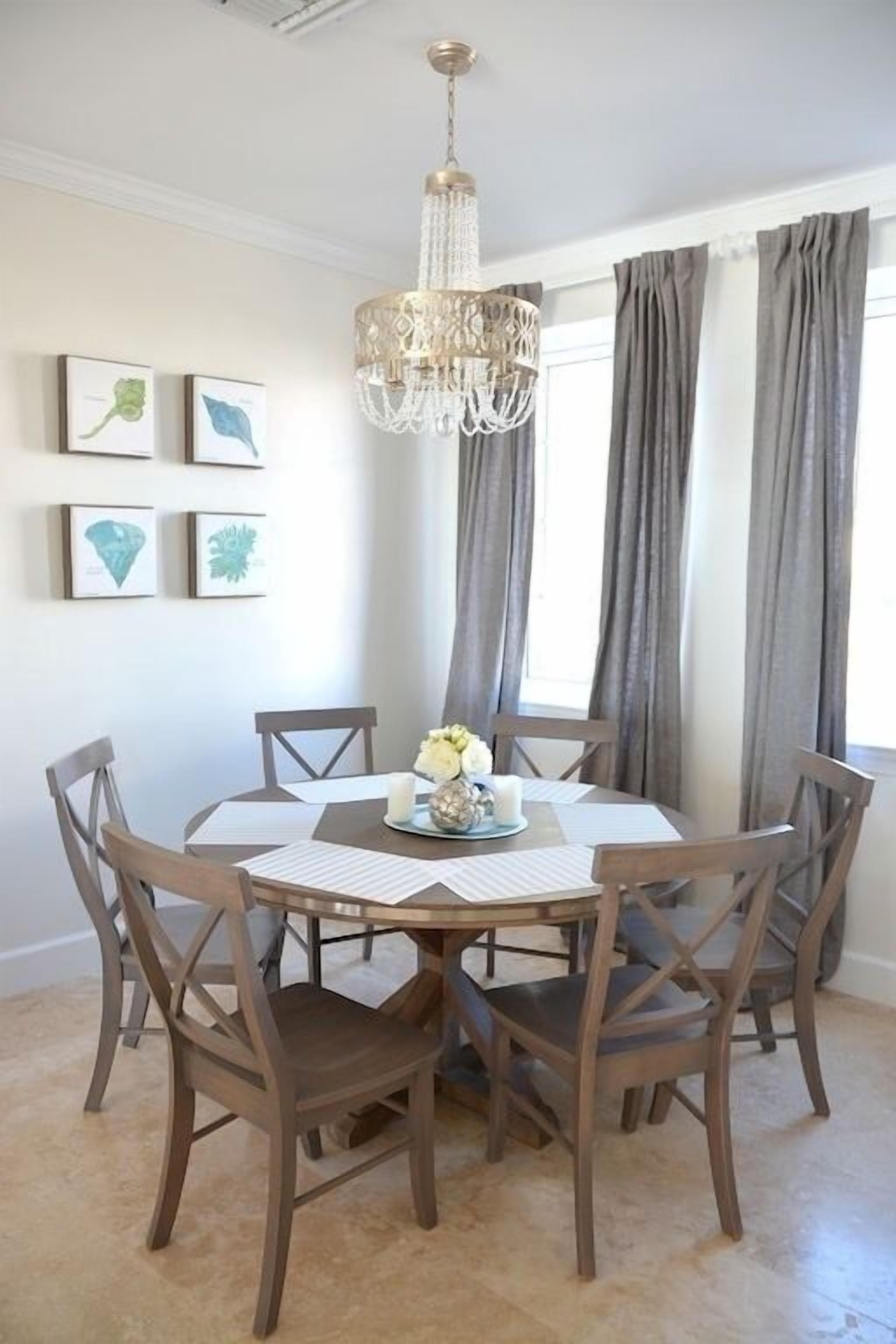 A dining area with a round table, six wooden chairs, a chandelier, and framed artwork on the wall.