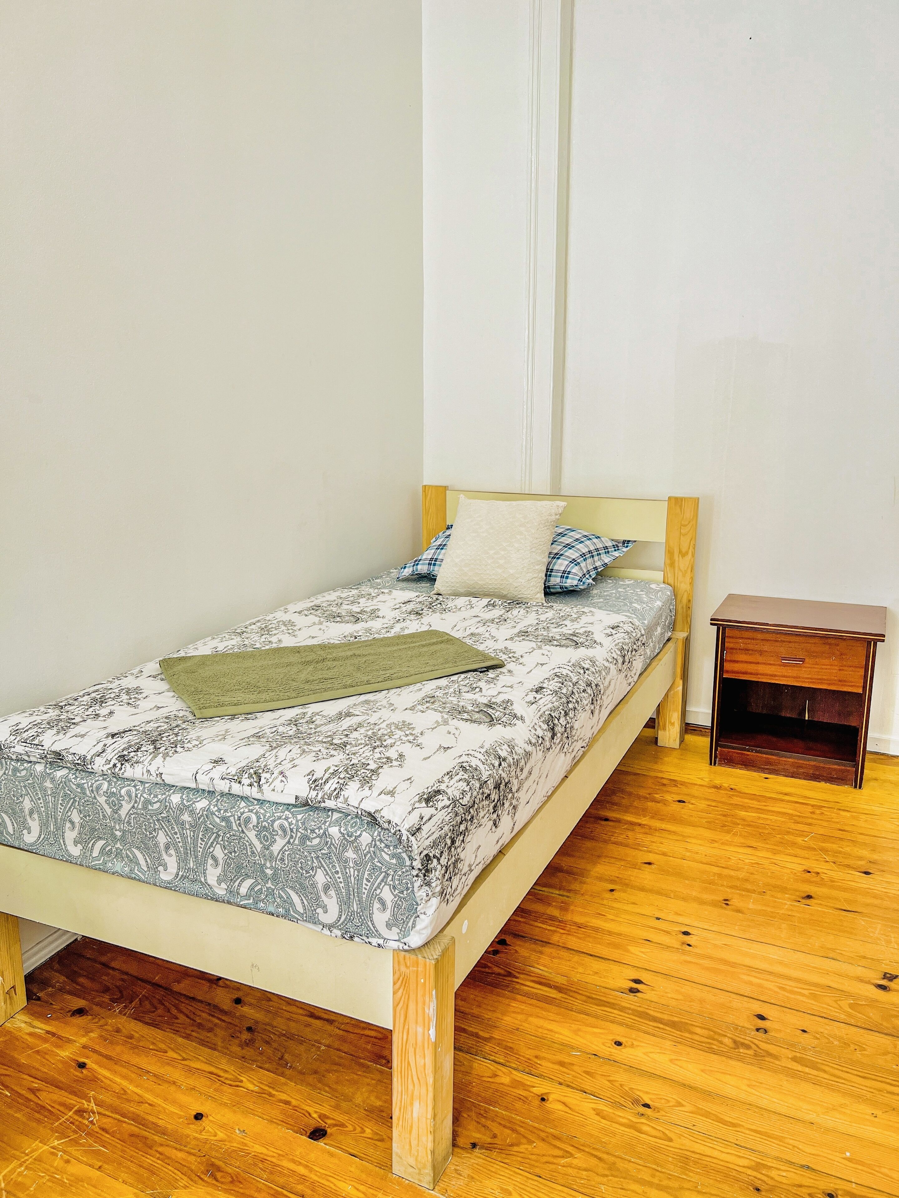 Una cama individual con un edredón estampado, una mesita de noche de madera y piso de madera.