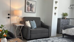 Una sala de estar moderna con un sofá gris, una mesita de noche con una lámpara, una obra de arte enmarcada, una alfombra gris y un aparador con una planta.