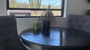 Un comedor con una mesa redonda, dos sillas tapizadas grises y una ventana que da a vista de un cactus y árboles.