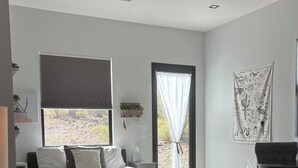 Un dormitorio moderno con una cama grande, un escritorio y una silla. Hay una ventana con persianas, una puerta con cortina y una estantería con plantas.