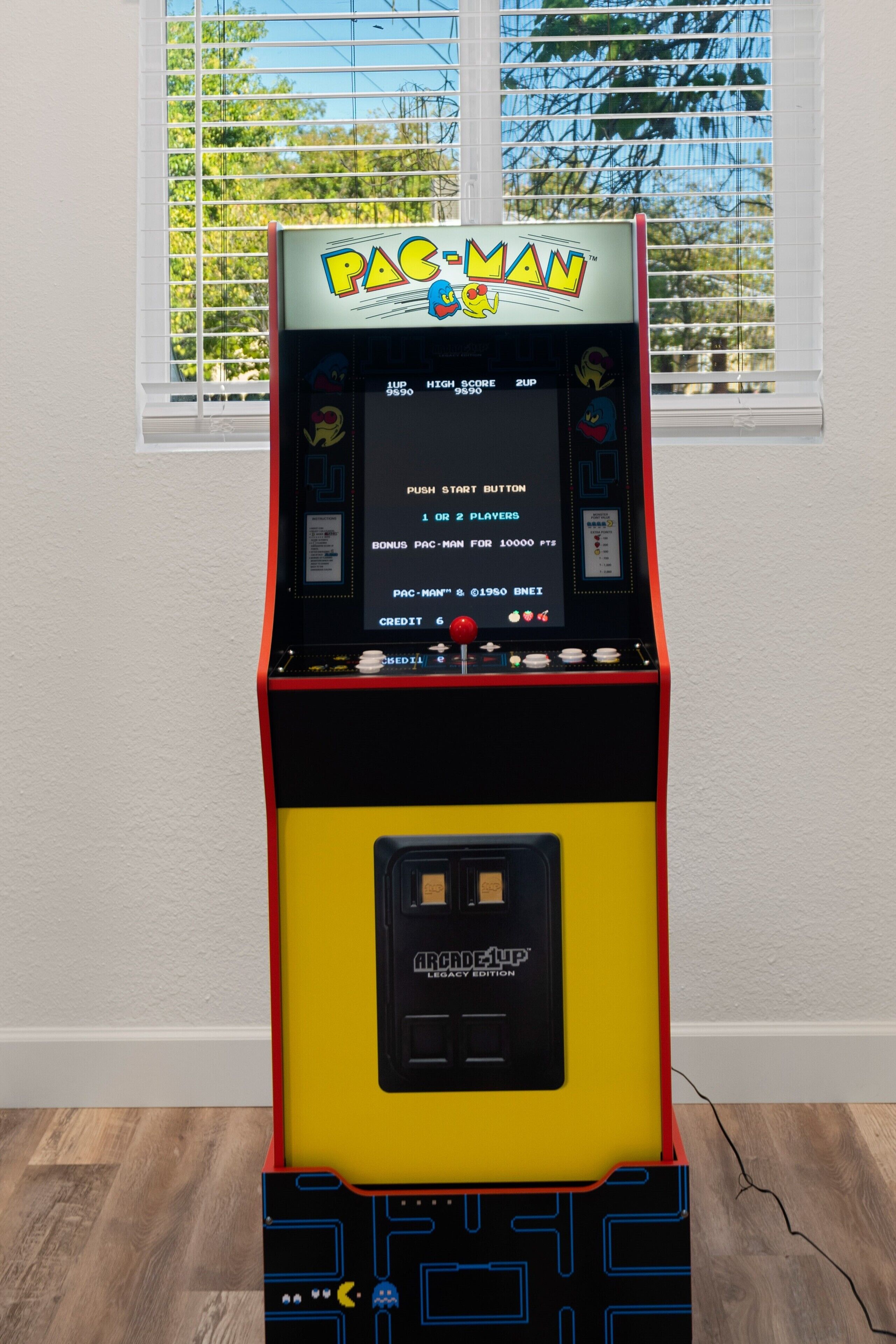 Une borne d’arcade Pac-Man dans une pièce avec une fenêtre et un plancher en bois.