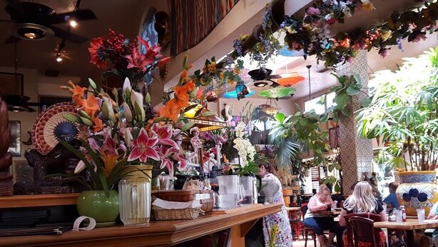 A restaurant with a variety of plants and flowers decorating the interior, wooden furniture, and a tiled floor.
