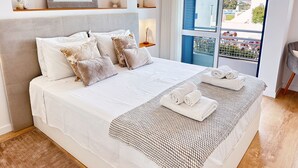 En pænt redt seng med hvide og beige puder, et vævet plaide og sammenrullet håndklæder på et hvidt sengetæppe.