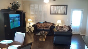 A living room with a dark wood floor, a television, a sofa, a coffee table, and a dining area with a table and chairs.