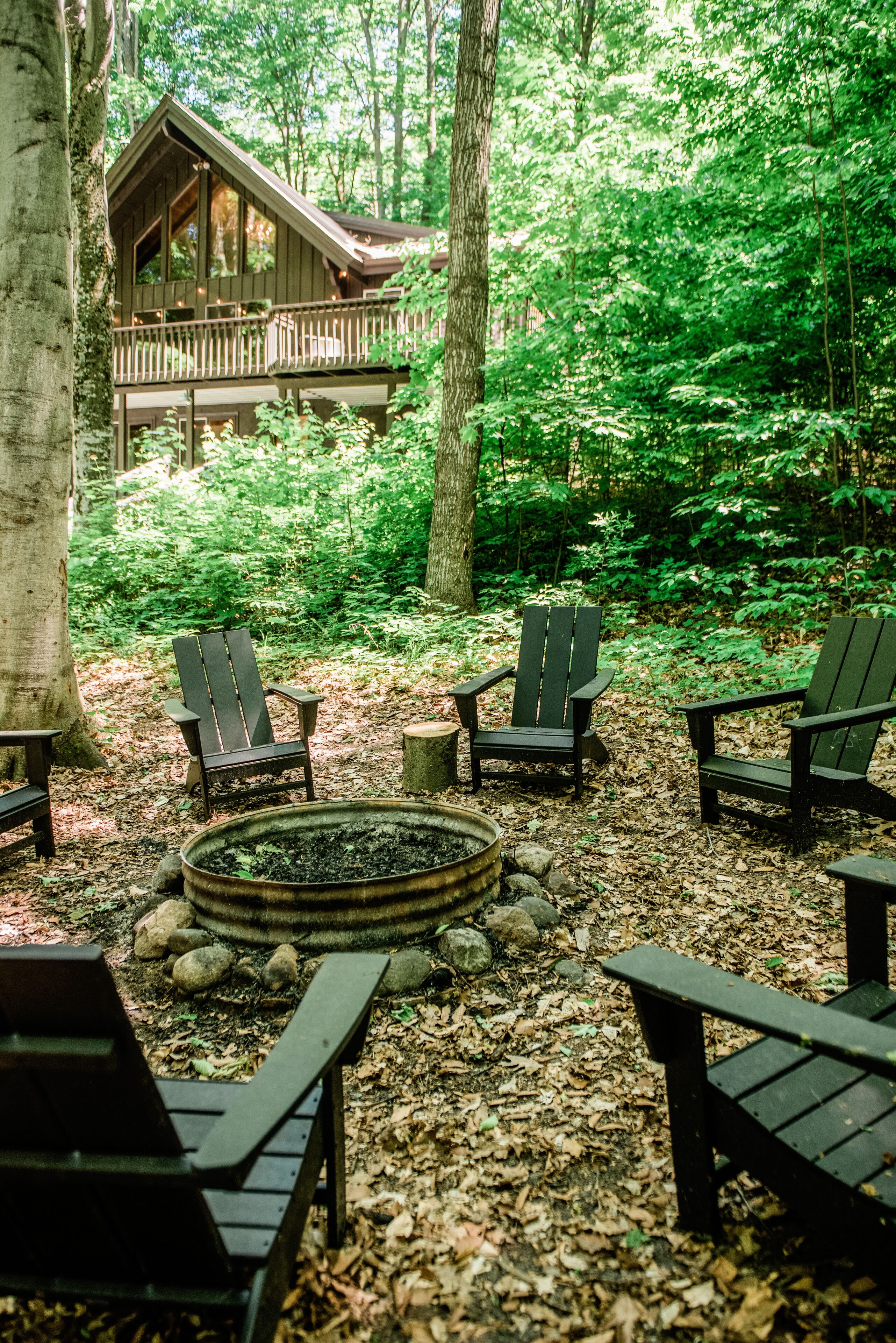Una cabaña con terraza, rodeada de bosque. Se ven sillas Adirondack y un brasero.