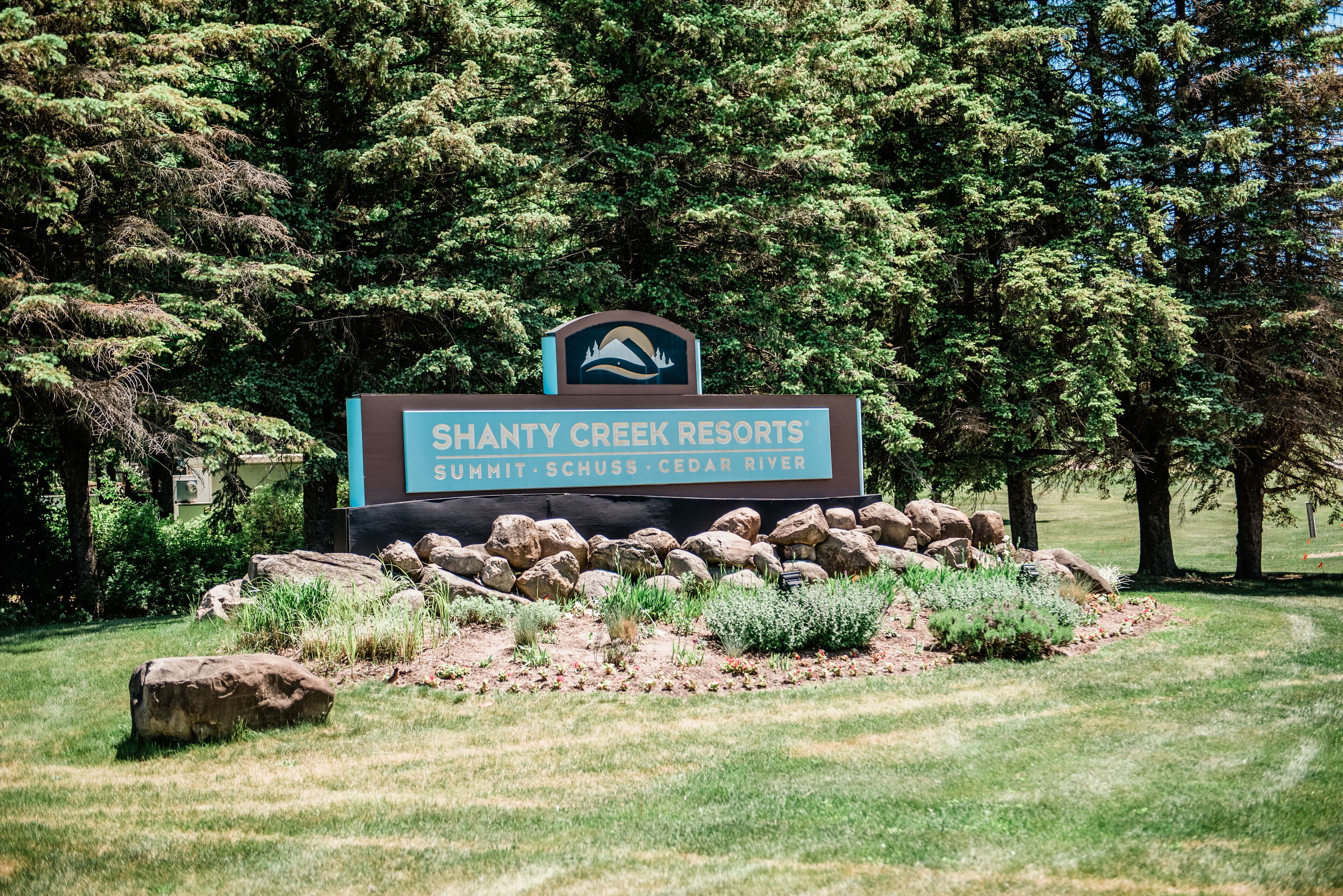 A sign for Shanty Creek Resorts with a mountainous logo, surrounded by rocks and greenery.