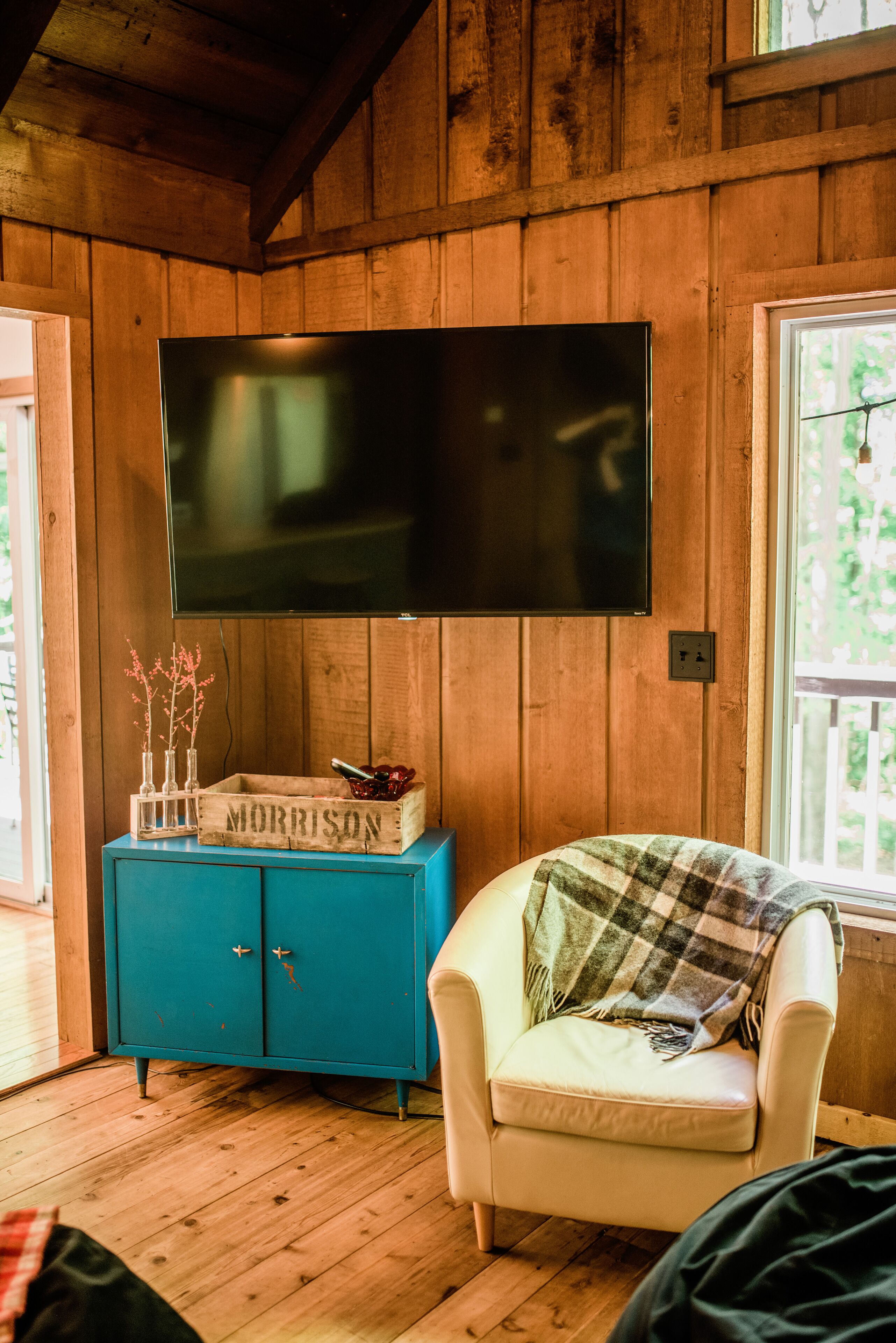 A wooden-paneled room with a mounted TV, a blue cabinet, and a plaid blanket on a white chair.