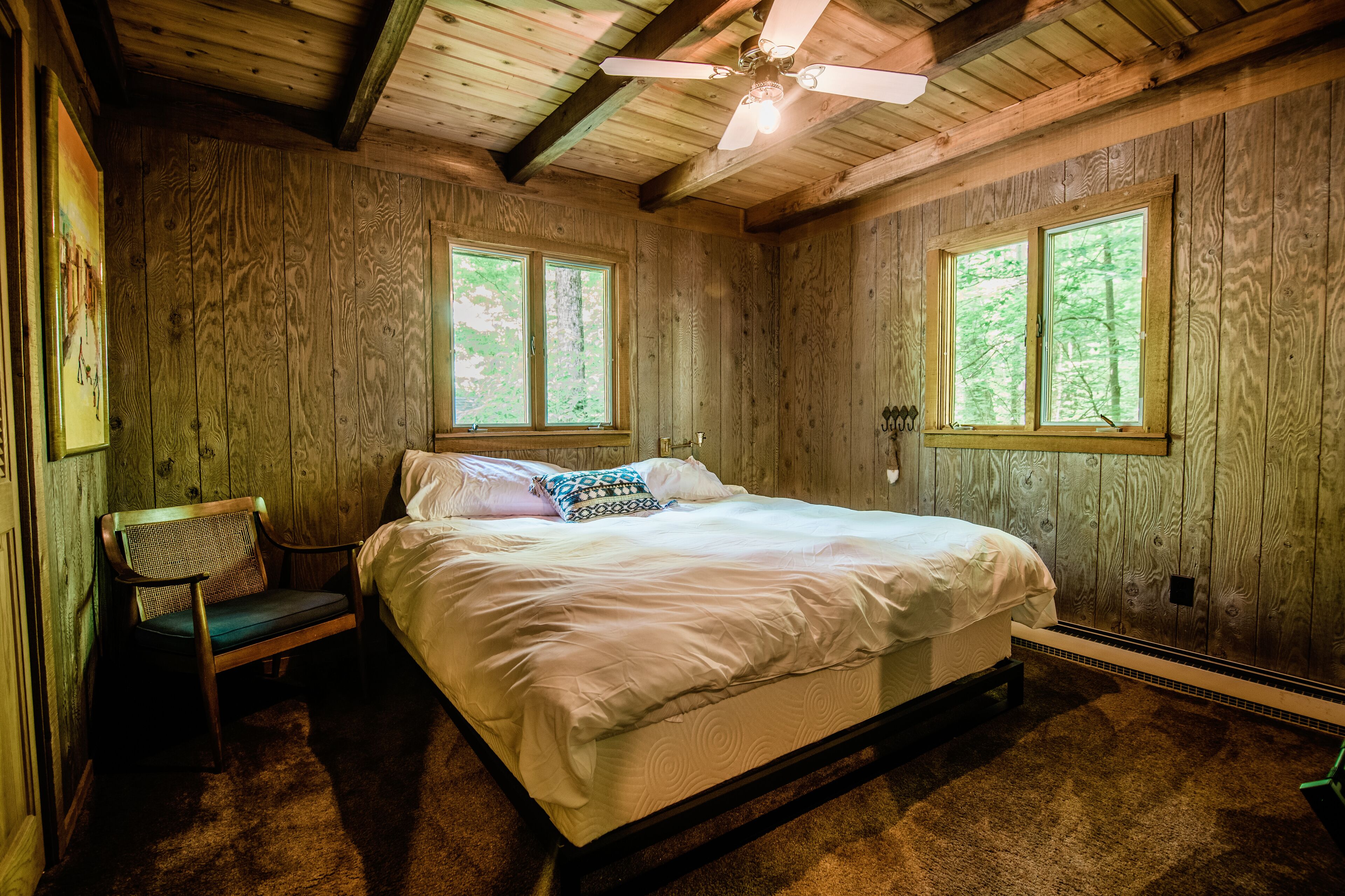 Slaapkamer met houten lambrisering, een bed, twee ramen, een plafondventilator en een bankje.