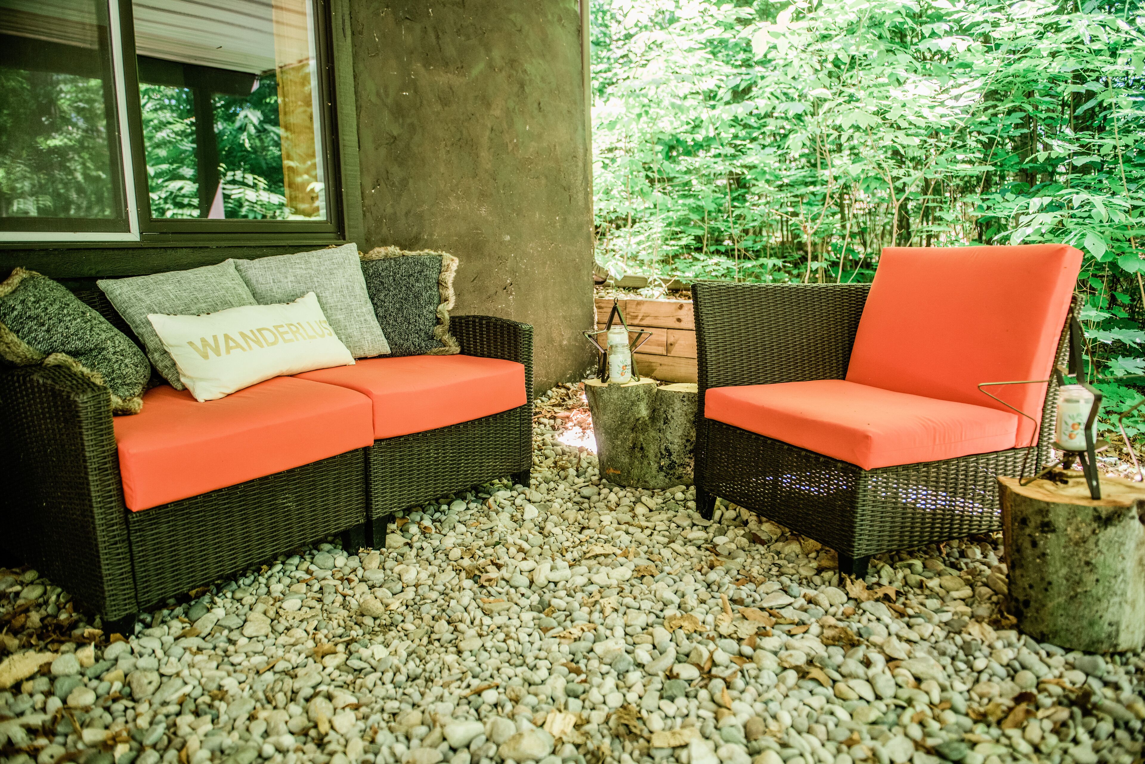 A wicker sofa with orange cushions and a cushion with "WANDERLUST" text, set on a gravel path with a wooden post nearby.