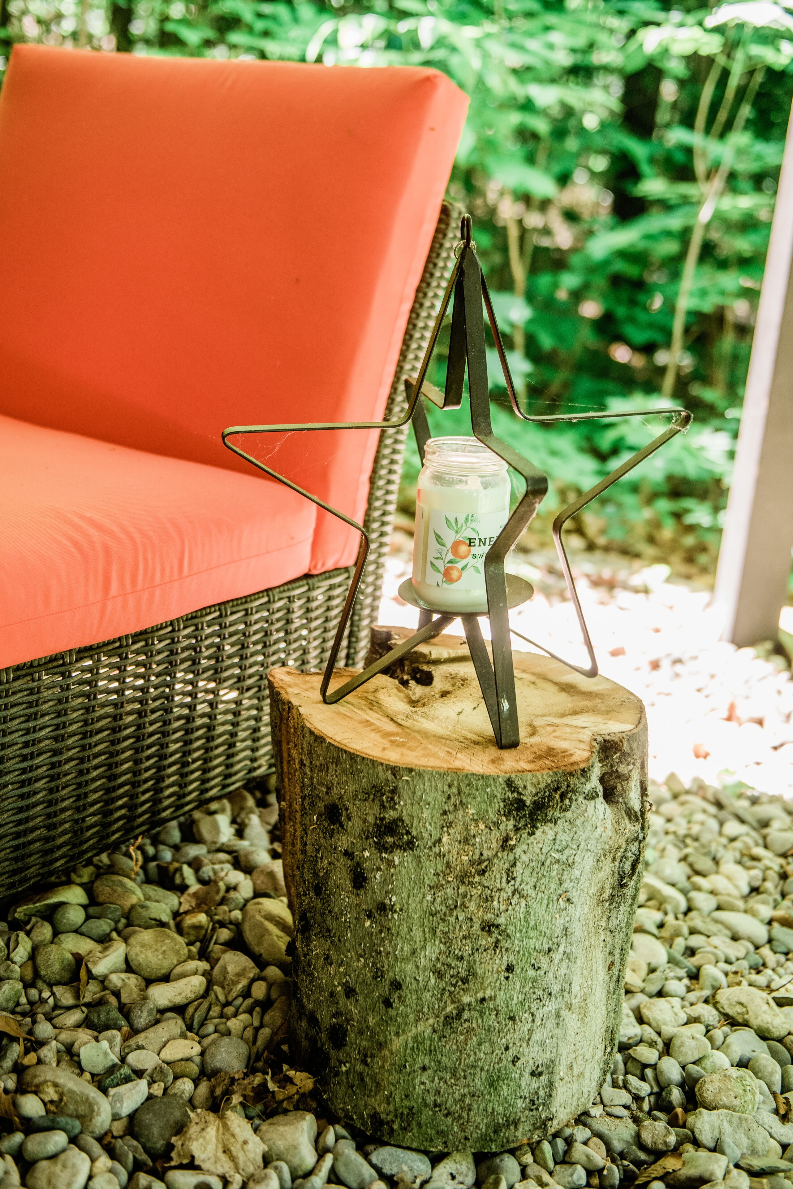 Une chaise en osier avec un coussin rouge, un chandelier posé sur une souche d’arbre, et un décor de jardin.