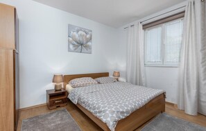 Ein Schlafzimmer mit einem hölzernen Bett, zwei Nachttischen mit Lampen, einem grauen Teppich, einem Fenster mit weißen Vorhängen und einem Blumenbild an der Wand.