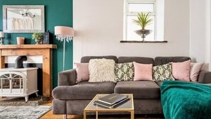 Een woonkamer met een bank, een klein bijzettafeltje, een zacht vloerkleed, een groene plaid en een raam met een plant.