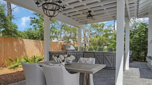 An outdoor patio with a wooden table and white cushioned chairs, a grill, and a pergola structure.