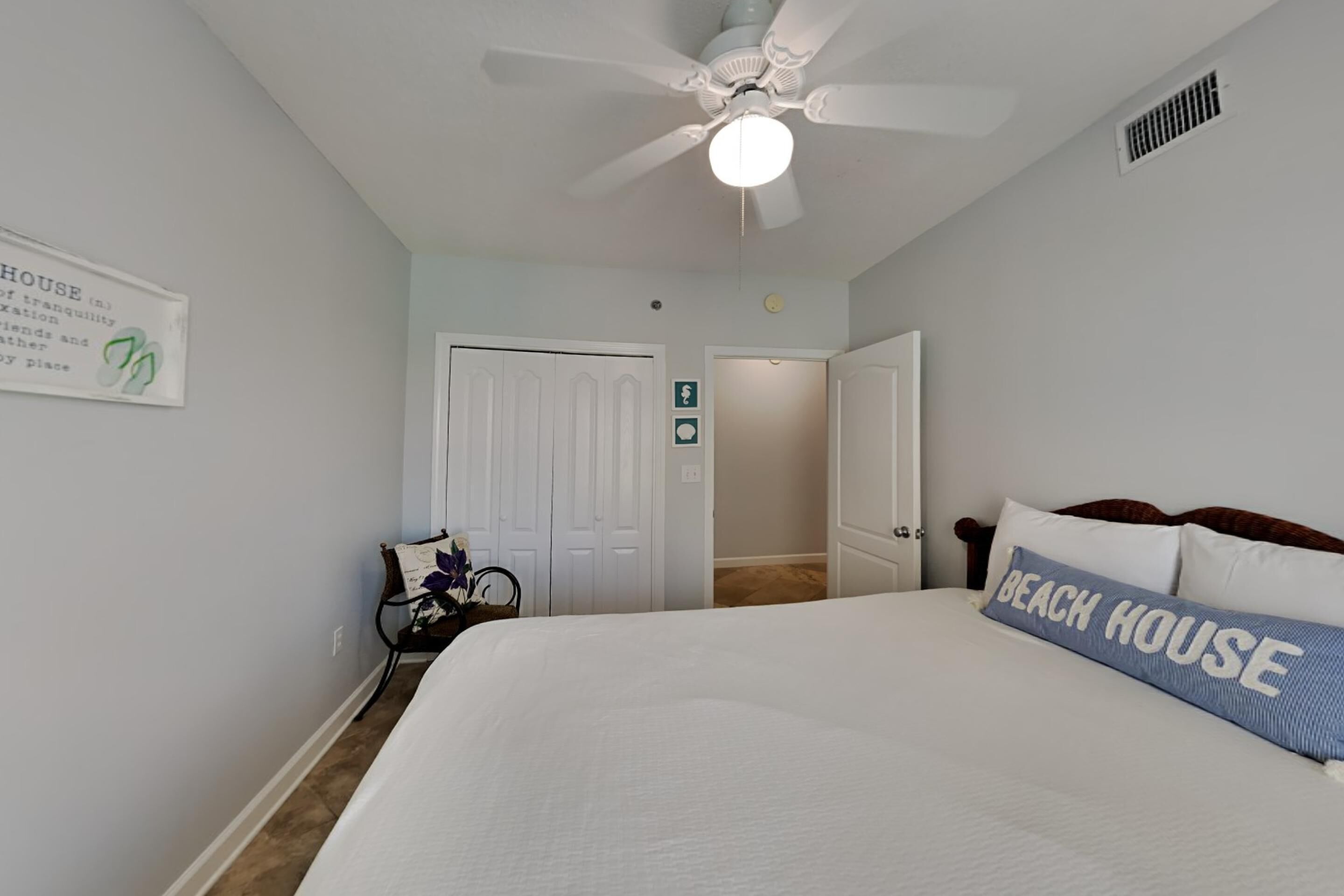 A bedroom with a bed, a ceiling fan, a sign on the wall, and a door leading to another room.