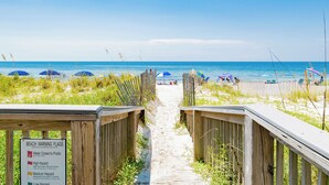 A beach pathway leading to the ocean with beach warning flags and umbrellas.