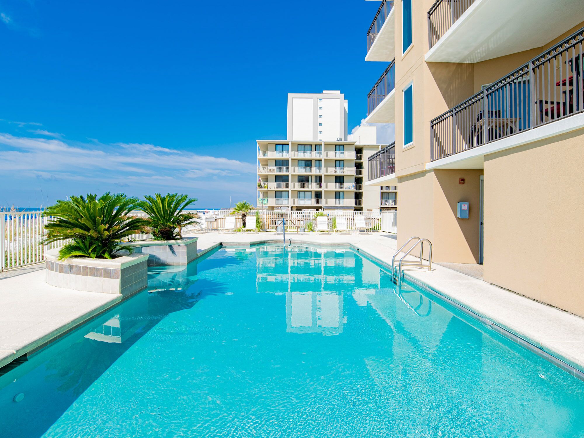 A swimming pool with a clear blue water, surrounded by a white fence, and a building with balconies in the background.