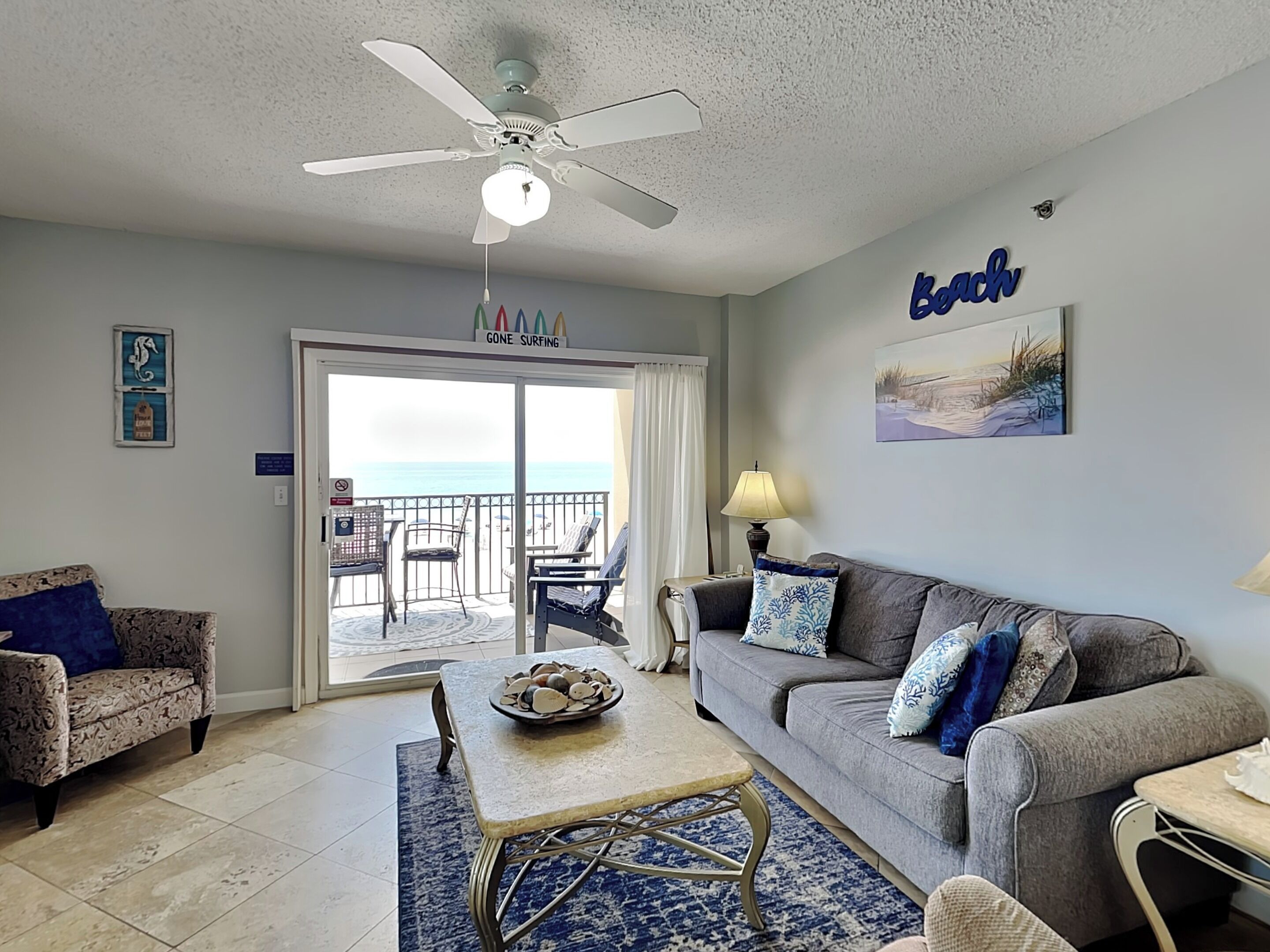 A living room with a ceiling fan, a couch, a coffee table, and a view of the ocean through a sliding glass door.
