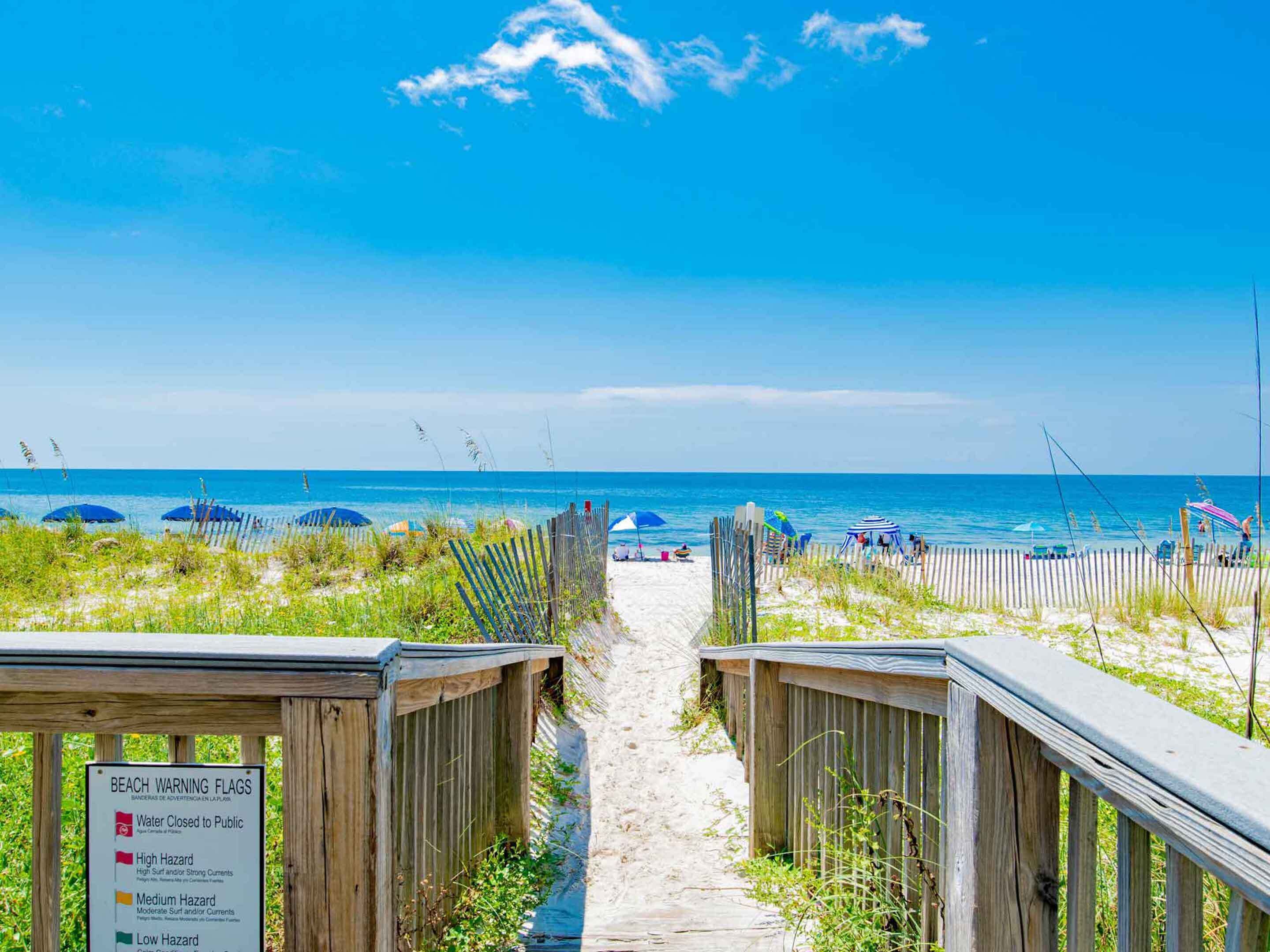 A beach path leading to the ocean with beach warning flags and umbrellas.