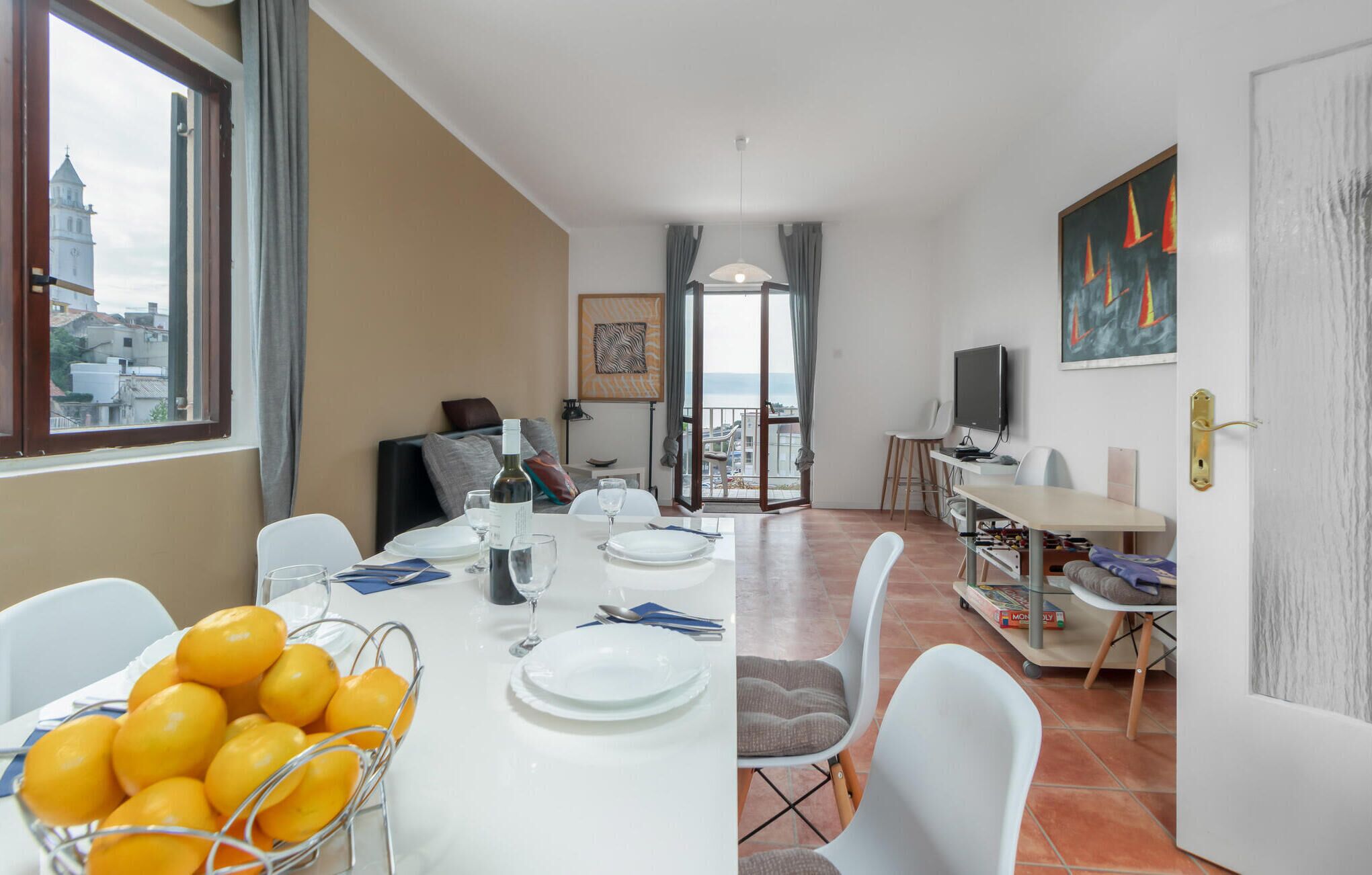 A dining area with a table set for two, a chair, a bowl of fruit, and a television in the background.