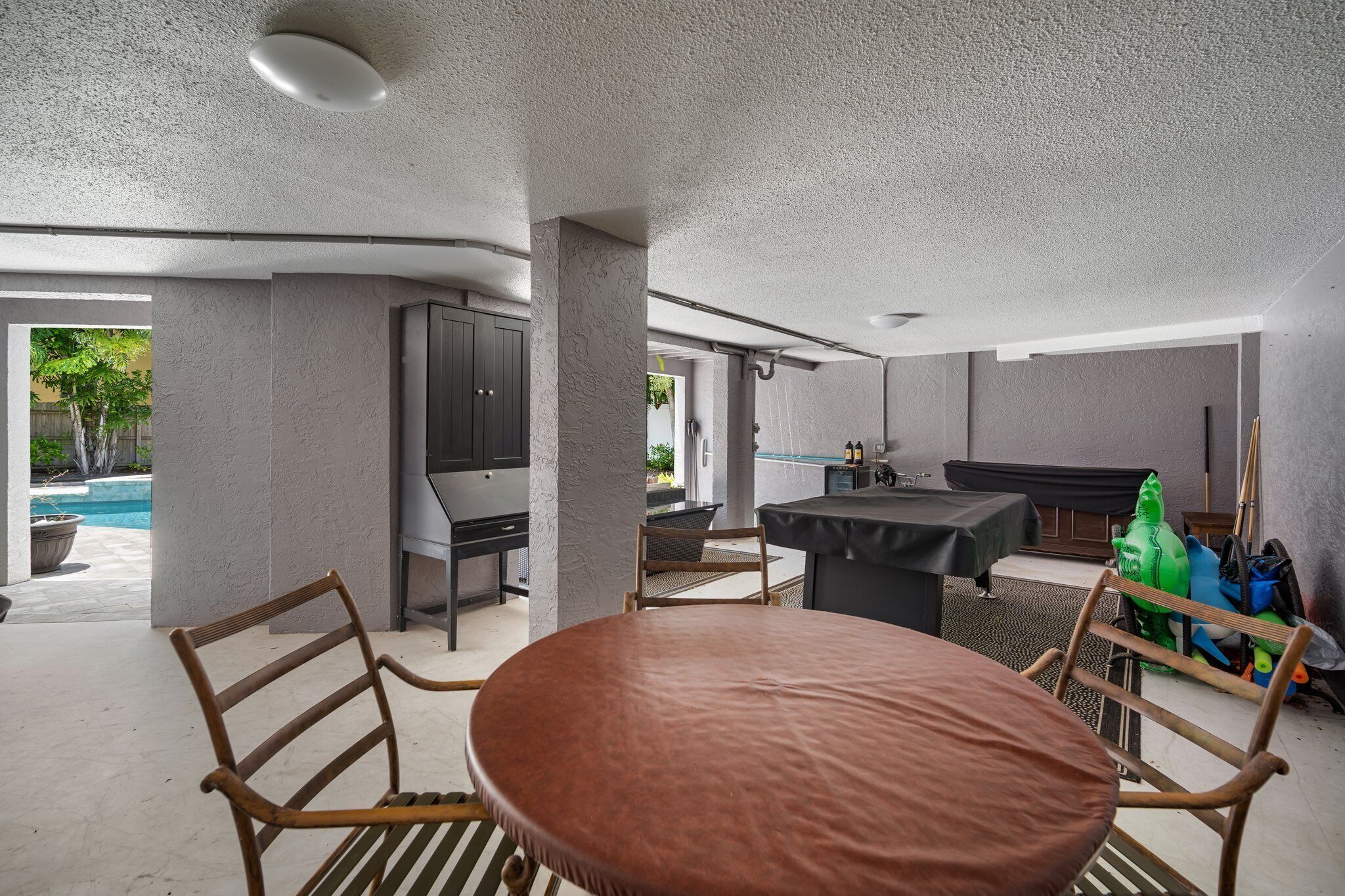 A modern indoor space with a round table and chairs, a pool visible through an open doorway, and a bar area with a grill.